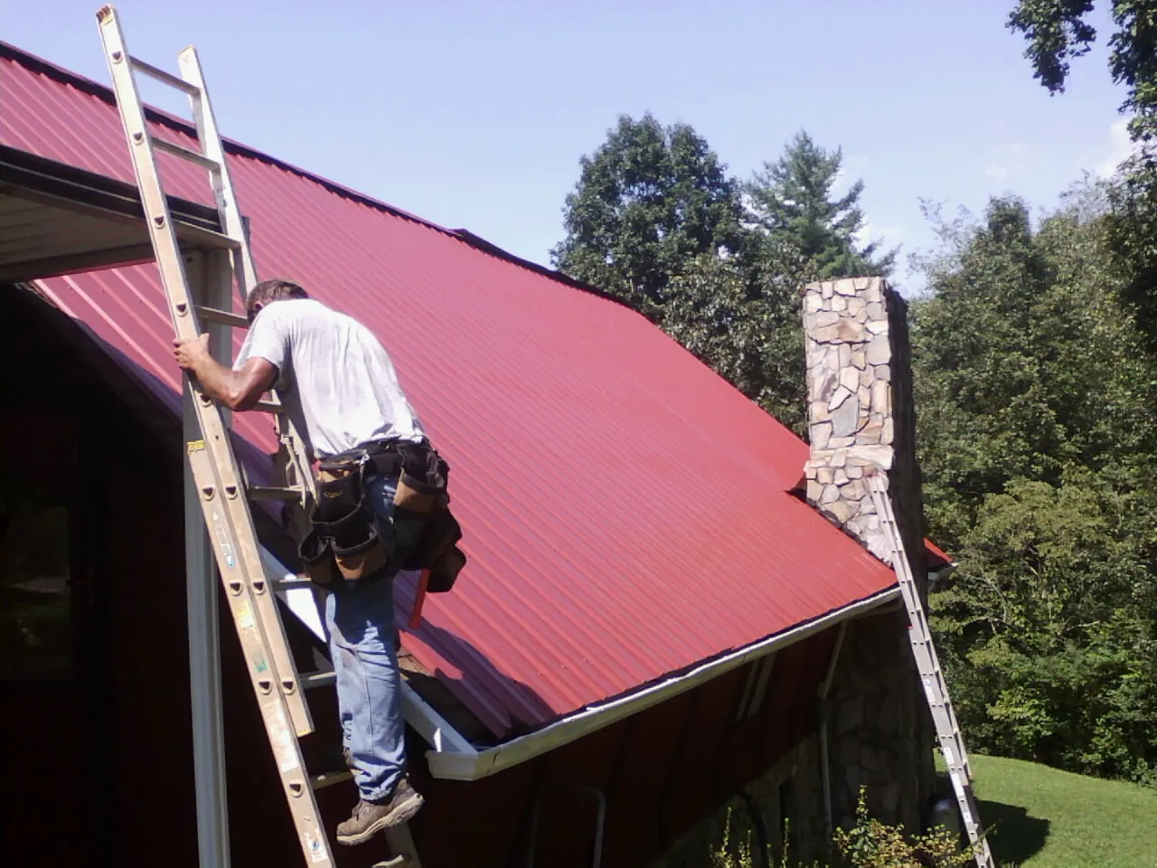 Roofer on a ladder beside a red metal roof and stone chimney — Soffit & Fascia Repair by Roofing Brothers in Chicago Ridge